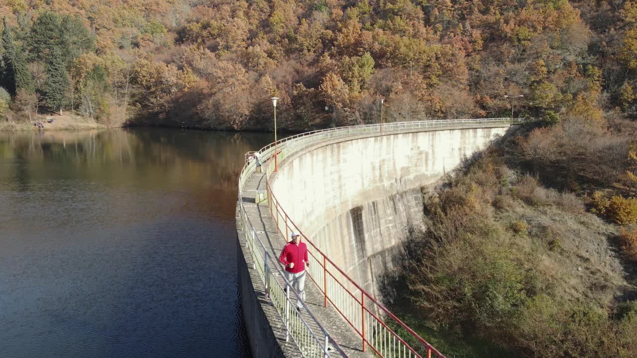 男子在大坝上慢跑，秋天的湖桥与山的彩色森林为背景，鸟瞰，海浪运动视频素材