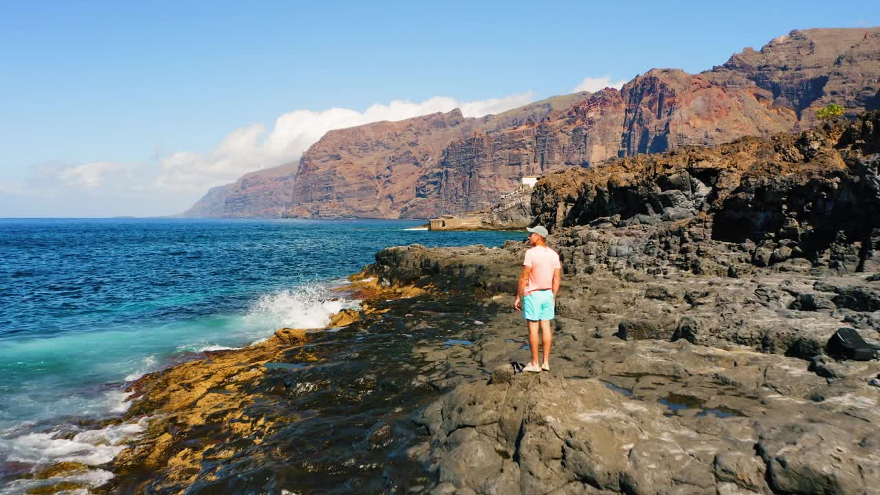 男子游客在火山特内里费岛的岩石海滩上欣赏海景。海浪溅起泡沫。巨人悬崖，阿坎提拉多斯·洛斯·吉甘特。暑假。空中无人机飞行。视频素材