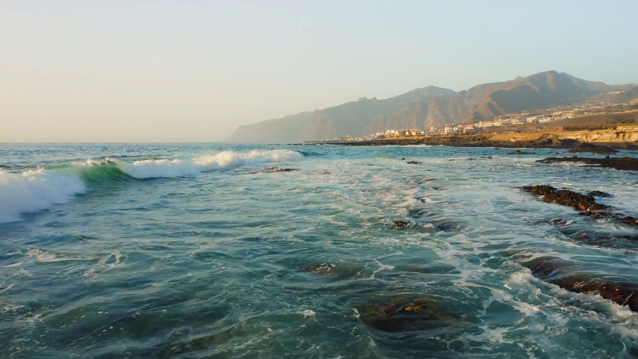 在暴风雨天气中飞越海浪表面。海浪泛起泡沫飞溅。背景是洛斯吉甘特悬崖的岩石海岸。美丽的景色碧蓝的大西洋水域。特内里费金丝雀。视频素材