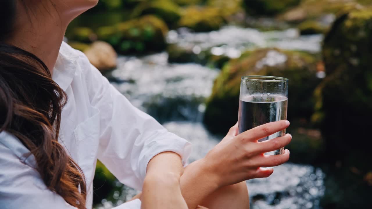 女人捧着一杯纯净的矿泉水在奔流的小溪边视频素材