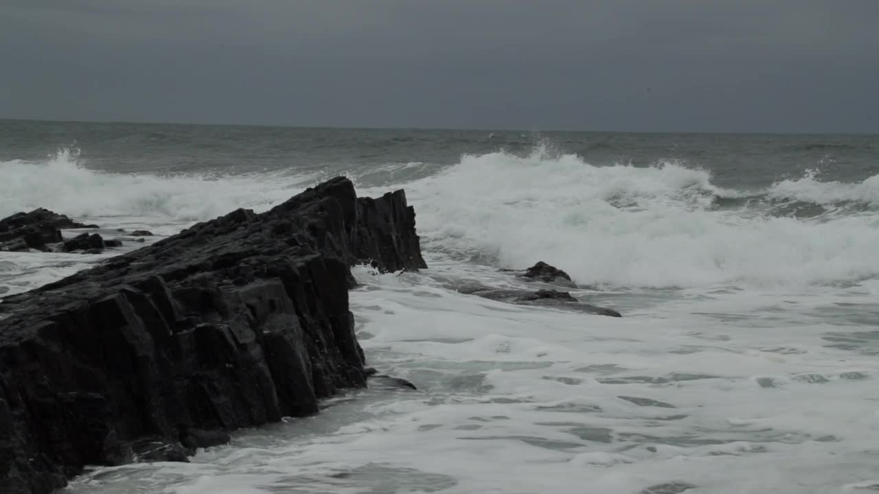 汹涌的海浪拍打着海滩上的黑色岩石，顶着暴风雨多云的天空视频素材