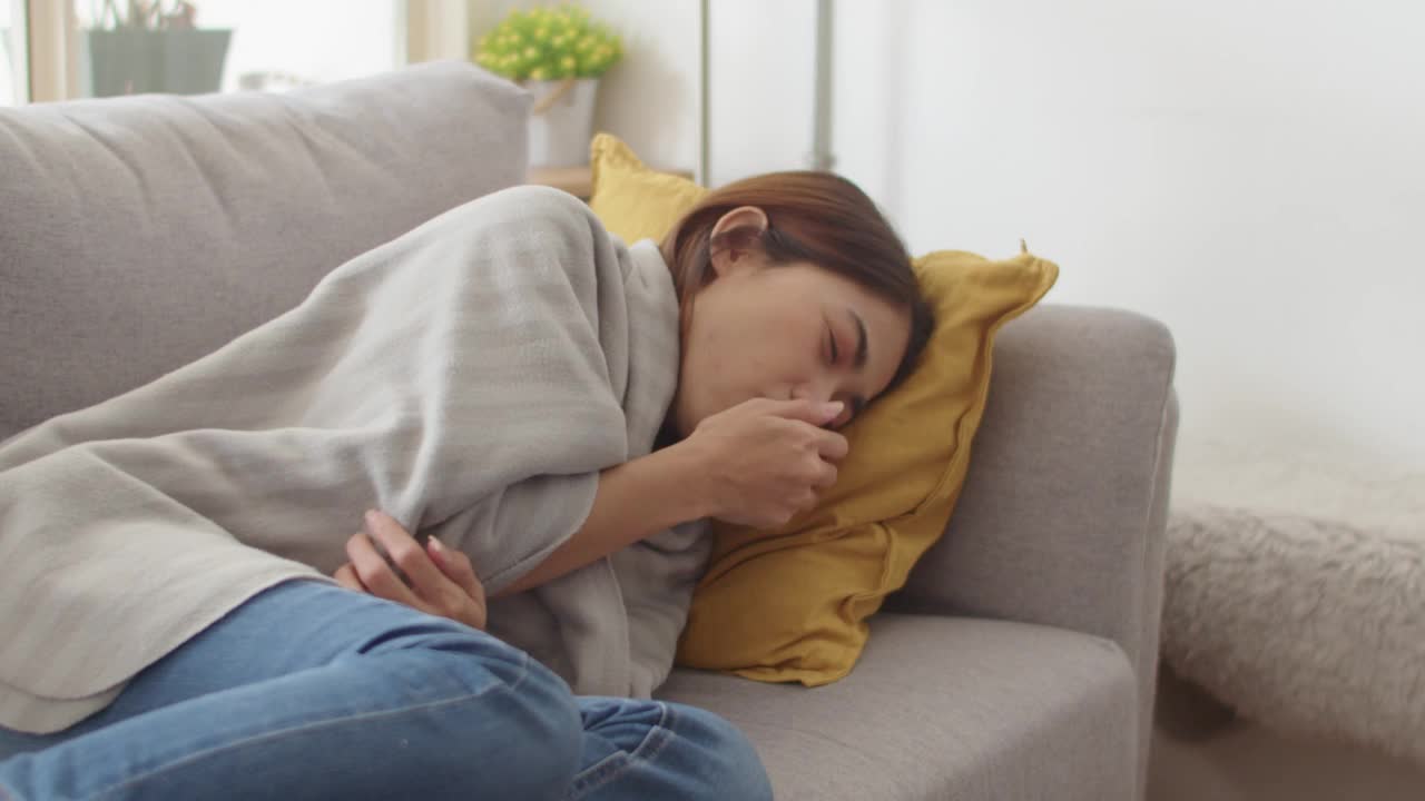 年轻的亚洲妇女坐在家里的沙发上感到恶心和打喷嚏。视频素材