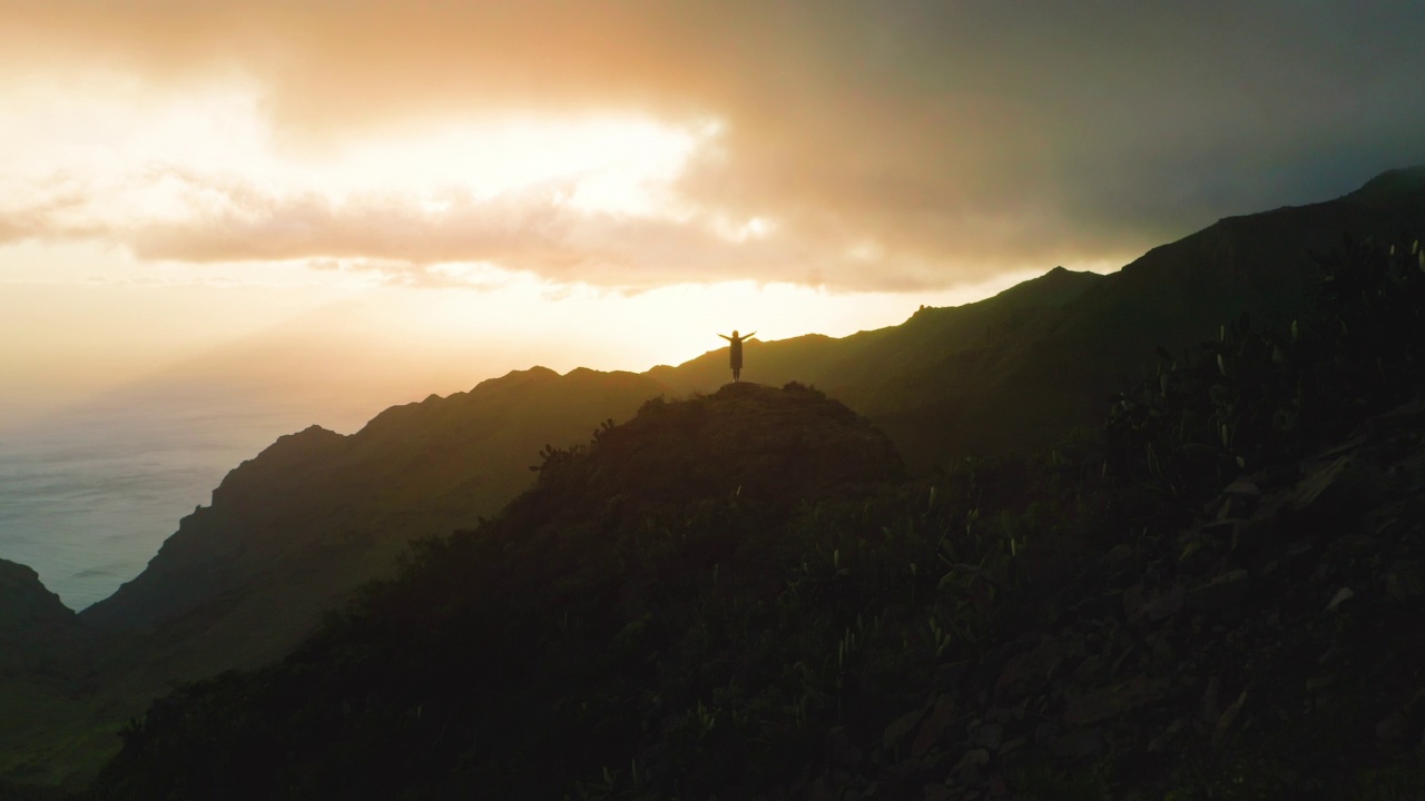 孤独的女孩站在岩石山顶，夕阳下的海洋背景。女人的剪影张开双臂，双手高举。天暗夜云。冬天。视频素材