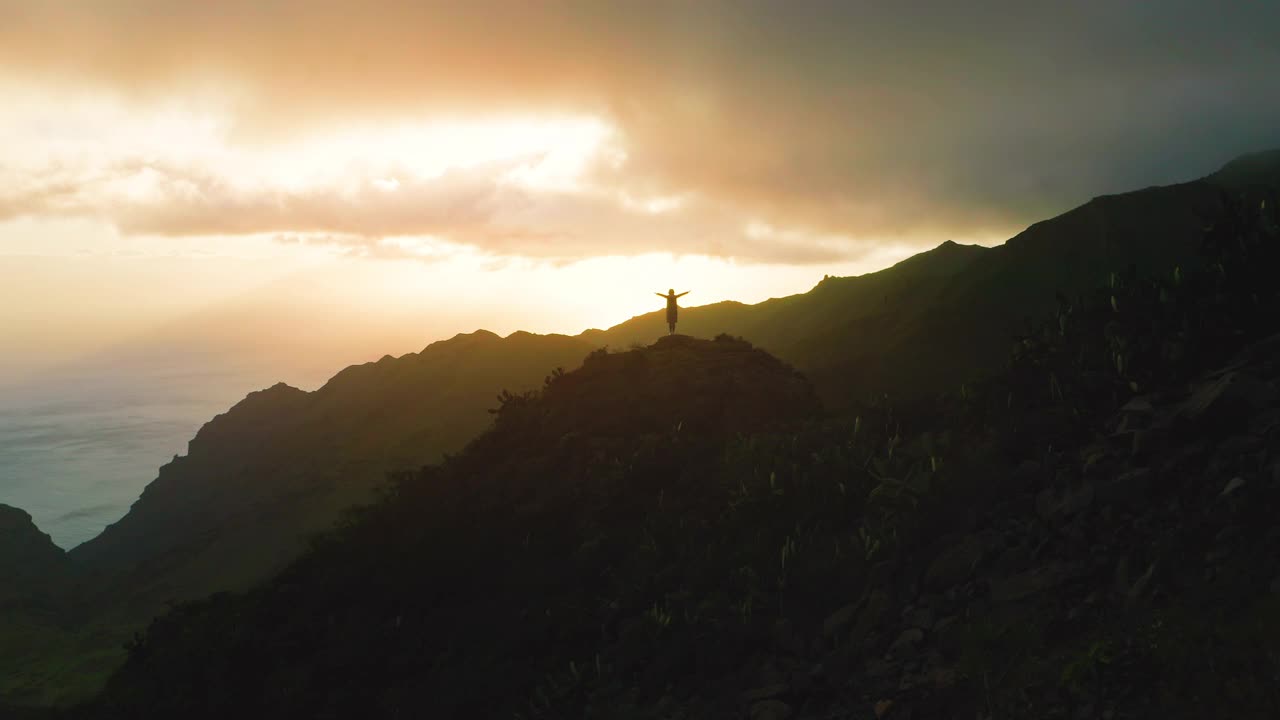 孤独的女孩站在岩石山顶，夕阳下的海洋背景。女人的剪影张开双臂，双手高举。天暗夜云。冬天。视频素材