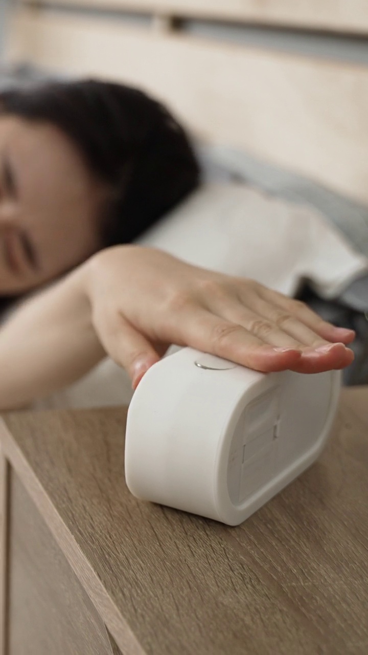 Vertical Screen: selective focus of a sleepy asian womanâs hand reaching out for the ringing alarm clock on nightstand and stopping it to sleep longer in the bedroom at home.视频素材