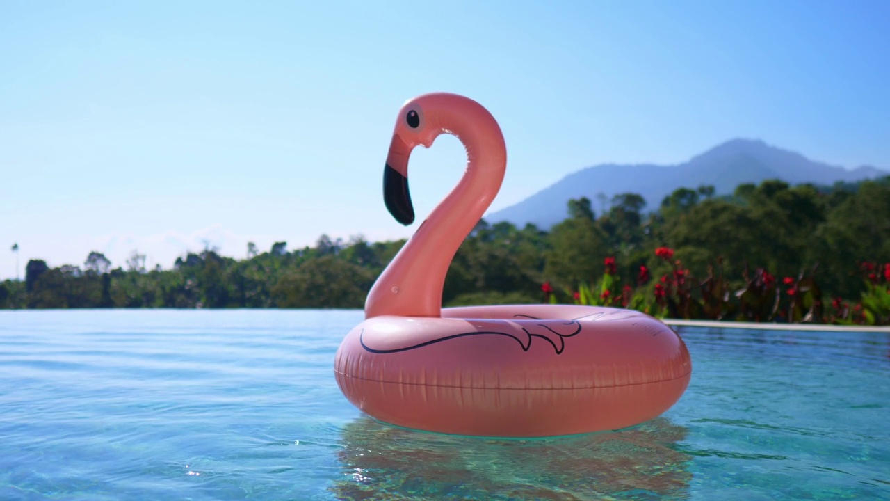 在一个阳光明媚的日子里，巨大的可充气的粉红色火烈鸟泳池漂浮玩具漂浮在游泳池里。在一个有充气火烈鸟游泳的游泳池的酒店休息。视频素材