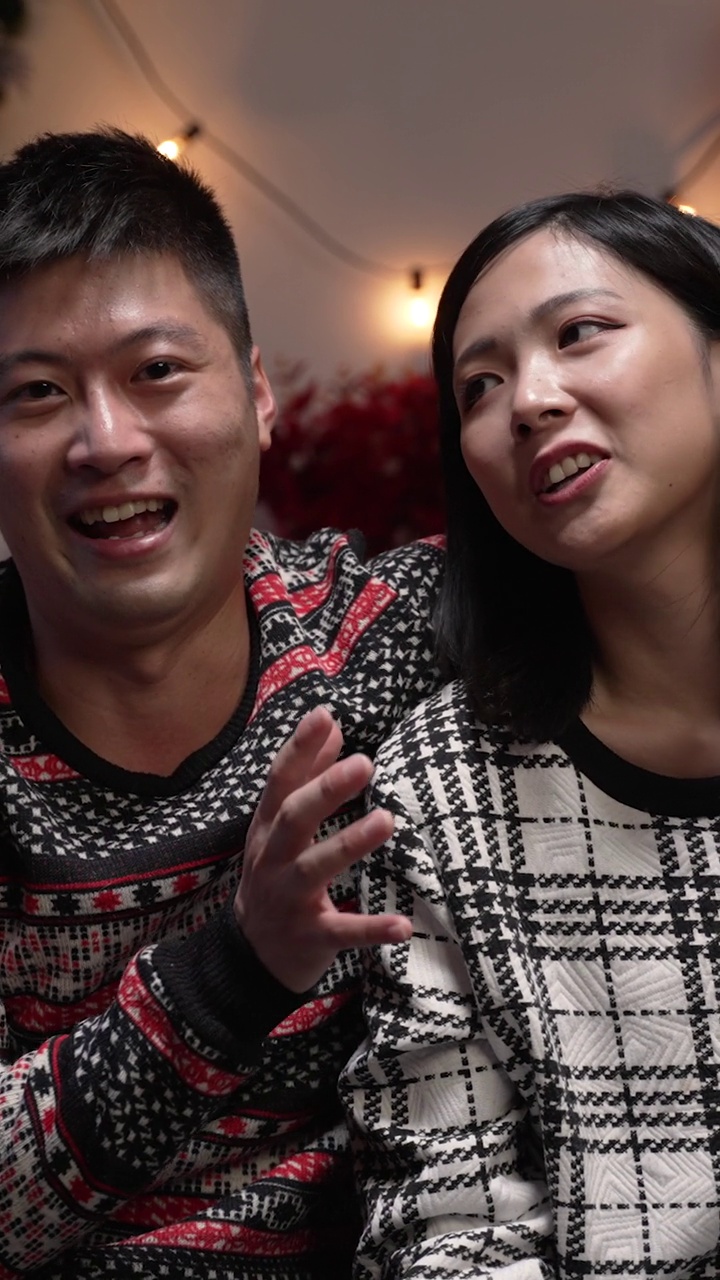 Vertical Screen: webcam view happy asian man talking with hand gestures with his arm around girlfriendâs shoulder whoâs holding his nose during a video call at home on Xmas eve视频素材