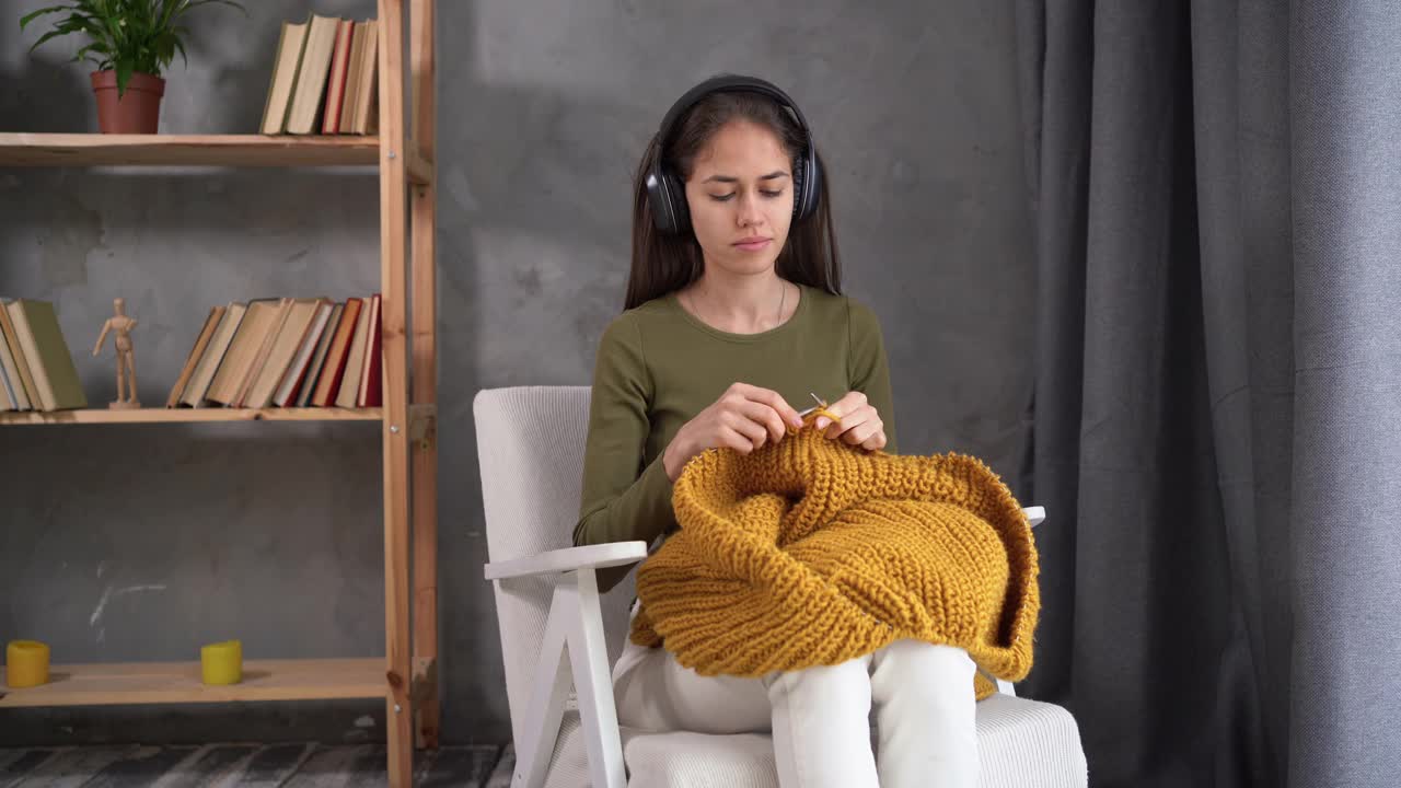 女人钩编。女孩坐在椅子上用毛线编织。我们用粗线编织。戴着耳机听音乐。假期的概念和周末爱好的职业。视频素材