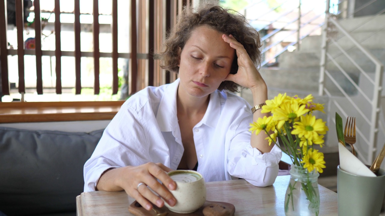 一位年轻沉思的白人女性独自坐在咖啡馆里，桌上放着一杯抹茶。她揉太阳穴以缓解头痛。心理健康概念视频素材