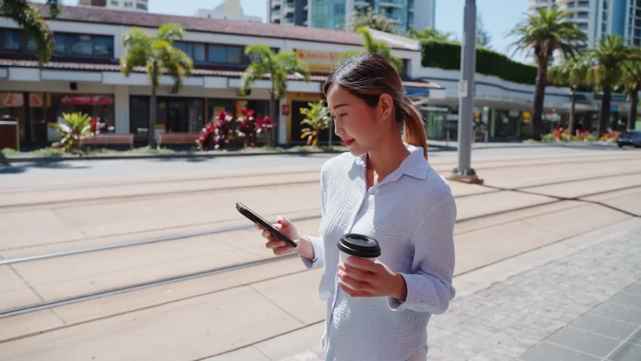 快乐的年轻中国女性拿着手机和咖啡，步行去市中心的办公室视频素材