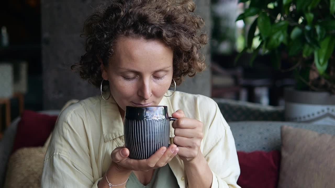 一位卷发美女在咖啡馆里吸着现煮咖啡的香气。女人端着一杯热腾腾的美国咖啡，享受着它的酸味，这让她充满活力。视频素材