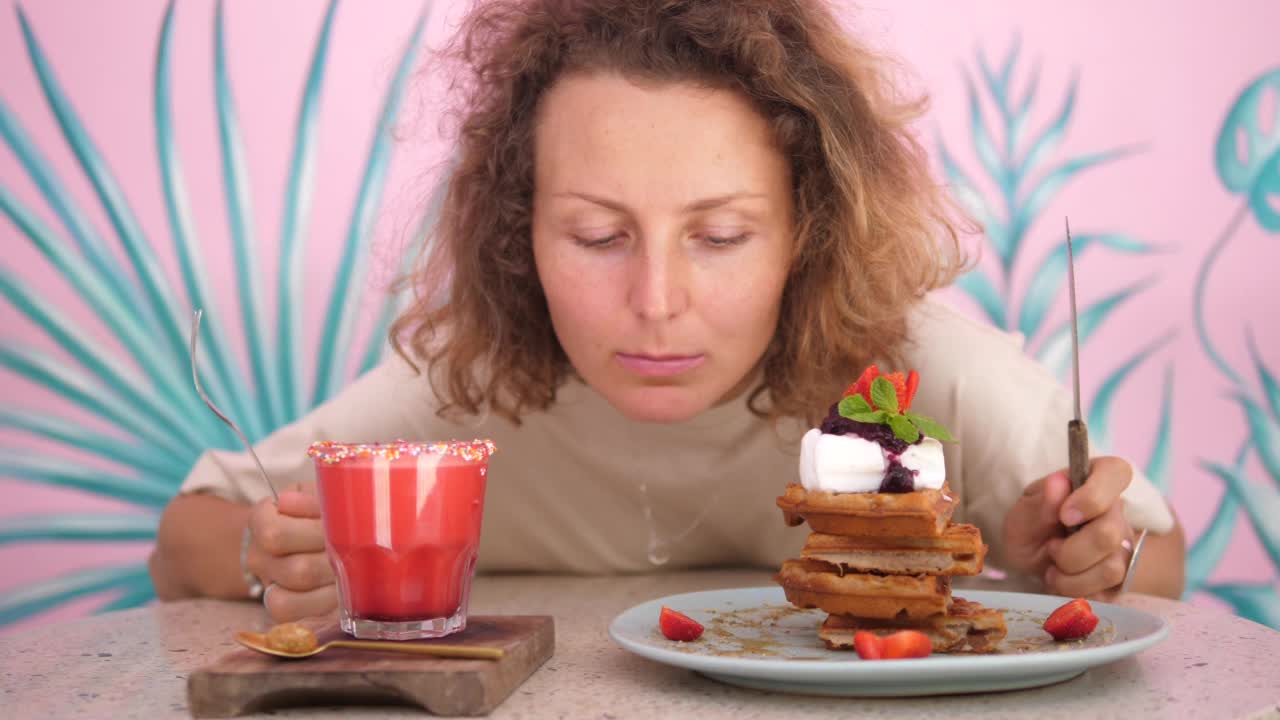 描绘了一个饥饿的白人妇女，她开玩笑地交替看看素食华夫饼，然后看看植物奶昔，期待着午餐。她决定不了从哪里开始吃饭。视频素材