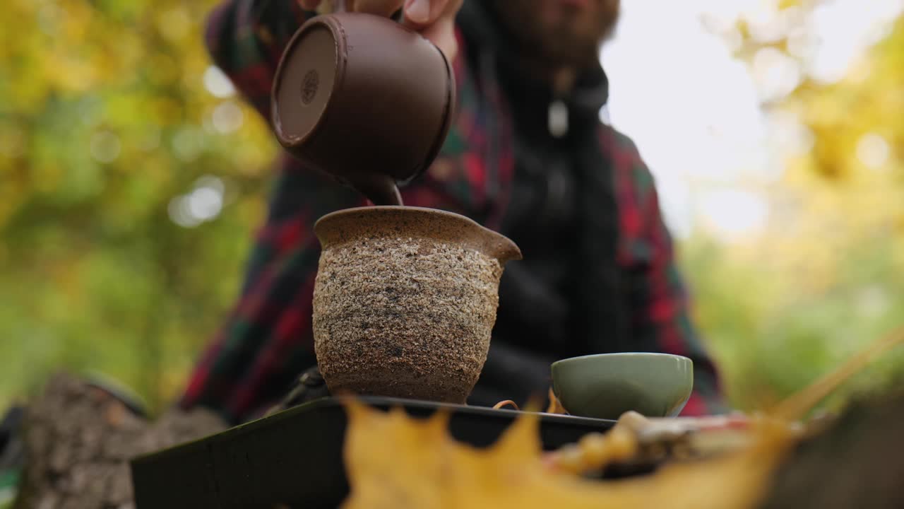 留胡子的茶艺师将泡好的普洱茶从茶壶中倒入陶壶中。户外茶道视频素材