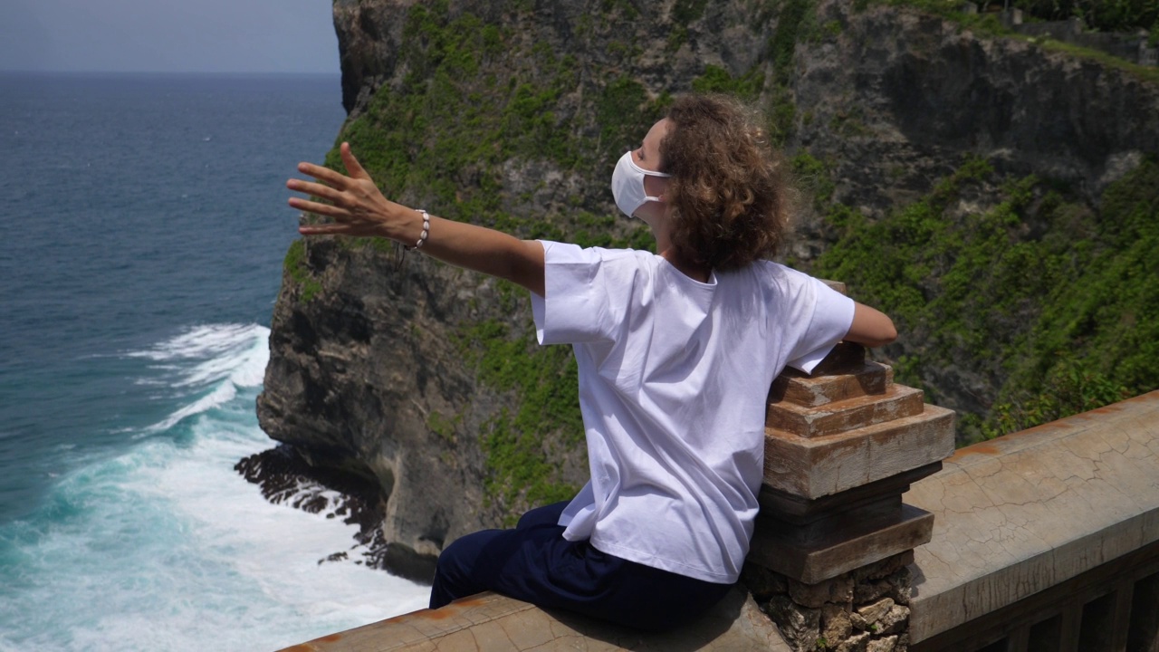 冠状病毒期间独自旅行。后视图的一个白人女孩卷头发戴着面具坐在阳台的边缘，她的双臂展开。视频素材