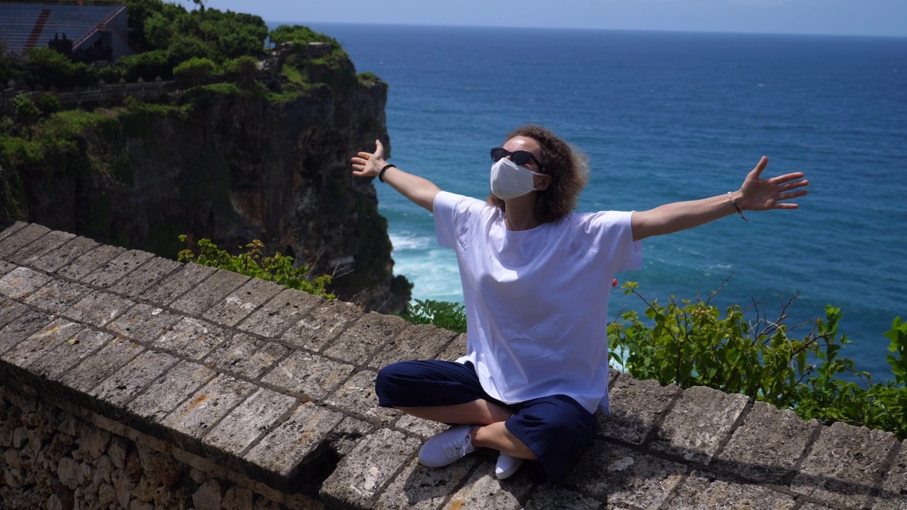 一名年轻的白人女性戴着口罩坐在观景台的阳台上，双臂张开。她坐在蓝色的大海前。冠状病毒期间旅行。视频素材