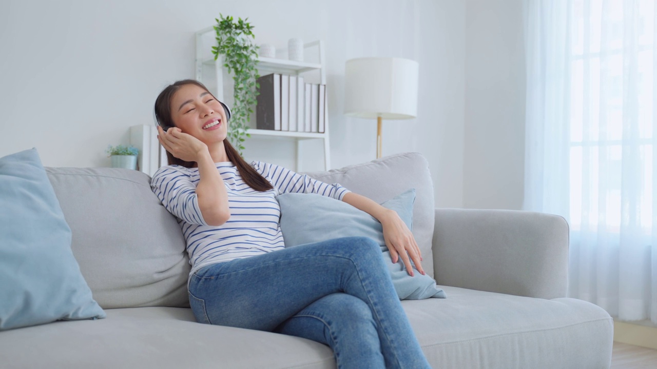 亚洲女人坐在沙发上听音乐跳舞。迷人美丽的年轻女孩戴上耳机，感觉很高兴在家里度过闲暇时间，在家里享受周末活动。视频素材