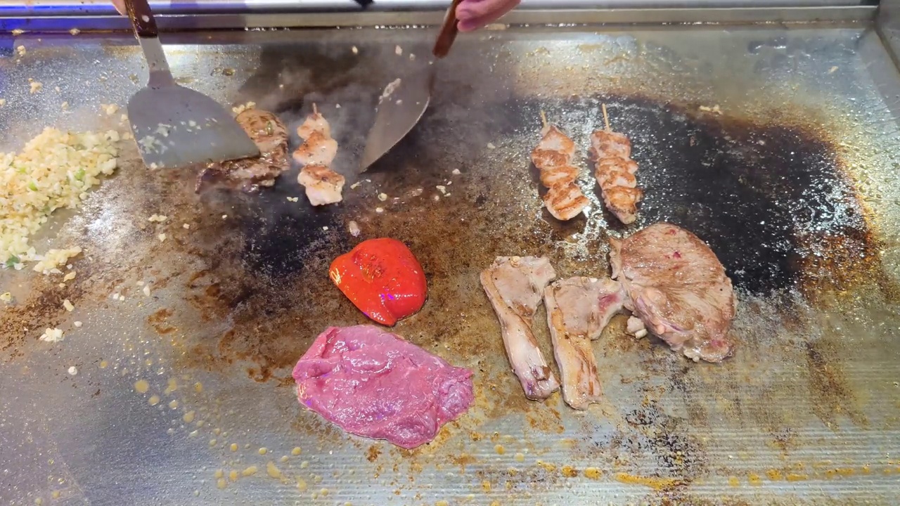 Tenderloin, steak, chicken skewers, peppers and lamb chops are topped with sliced ​​garlic and spring onions by an unrecognizable chef and then fried on a teriyaki grill plate in a restaurant视频素材