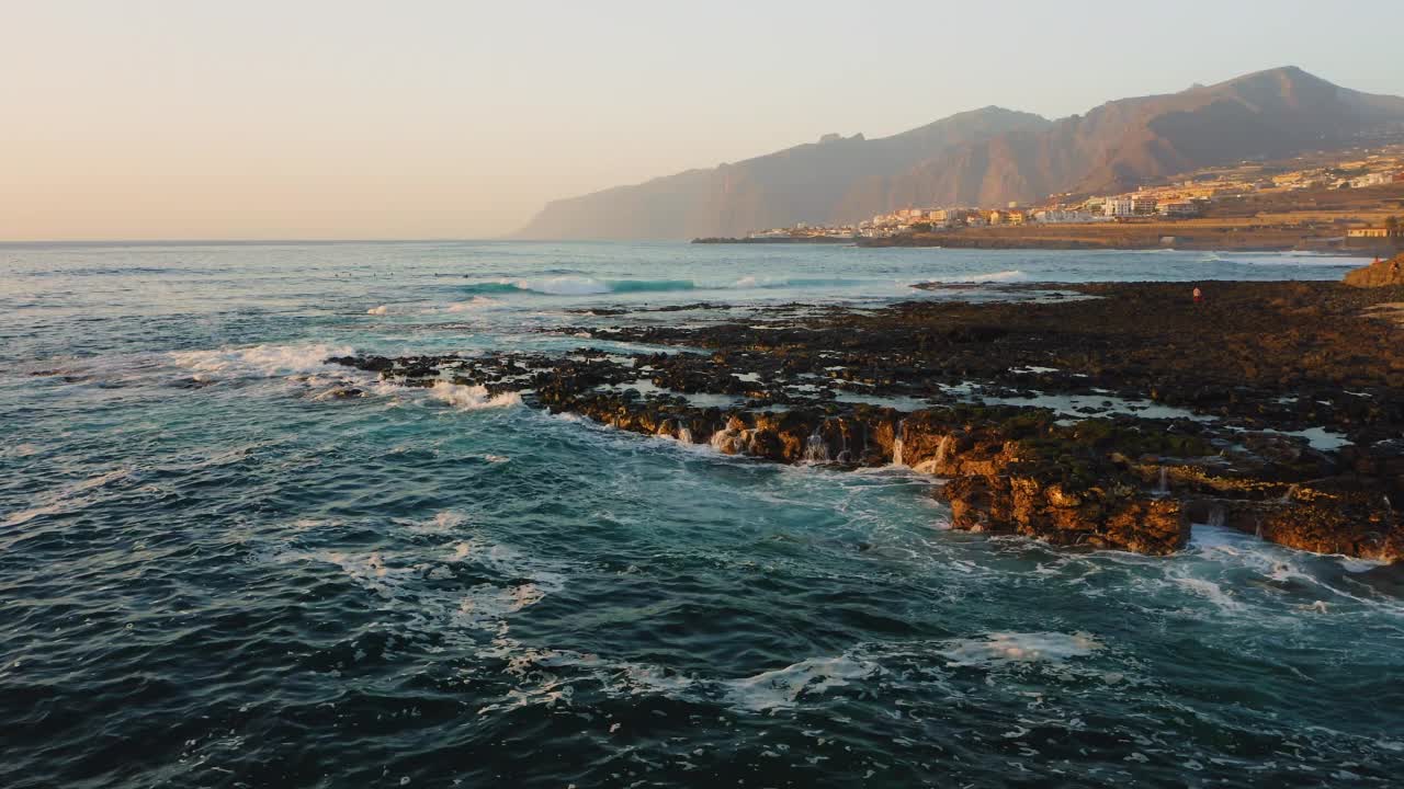 海浪撞击着度假小镇附近的特内里费加那利岛的岩石火山海滩。水沫飞溅。蓝蓝的天空下，陡峭的山崖被薄雾笼罩。空中镜头。视频素材