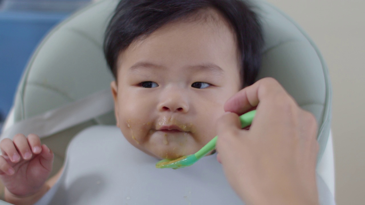 坐在高脚椅上的亚洲小男孩正在被妈妈喂食物。视频素材