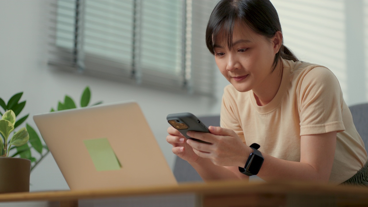 亚洲女性开心地微笑着坐在客厅的沙发上，用智能手机搜索网上购物或浏览社交媒体。视频素材