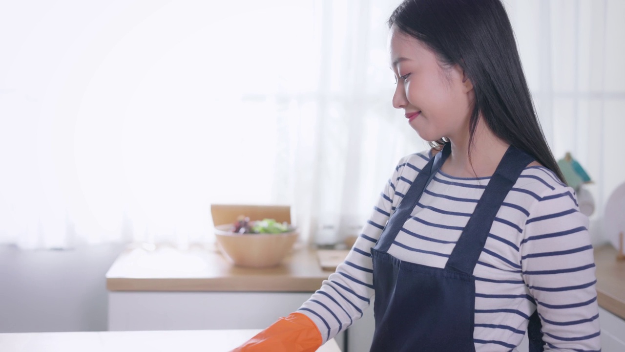 年轻的亚洲女子看着相机，用掸子打扫家。美丽的清洁服务人员打扫卫生和整理厨房。家务和杂务概念视频素材