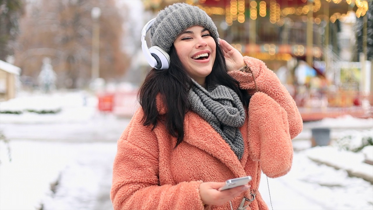 快乐的年轻女子在露天圣诞市场上戴着耳机听音乐跳舞视频下载