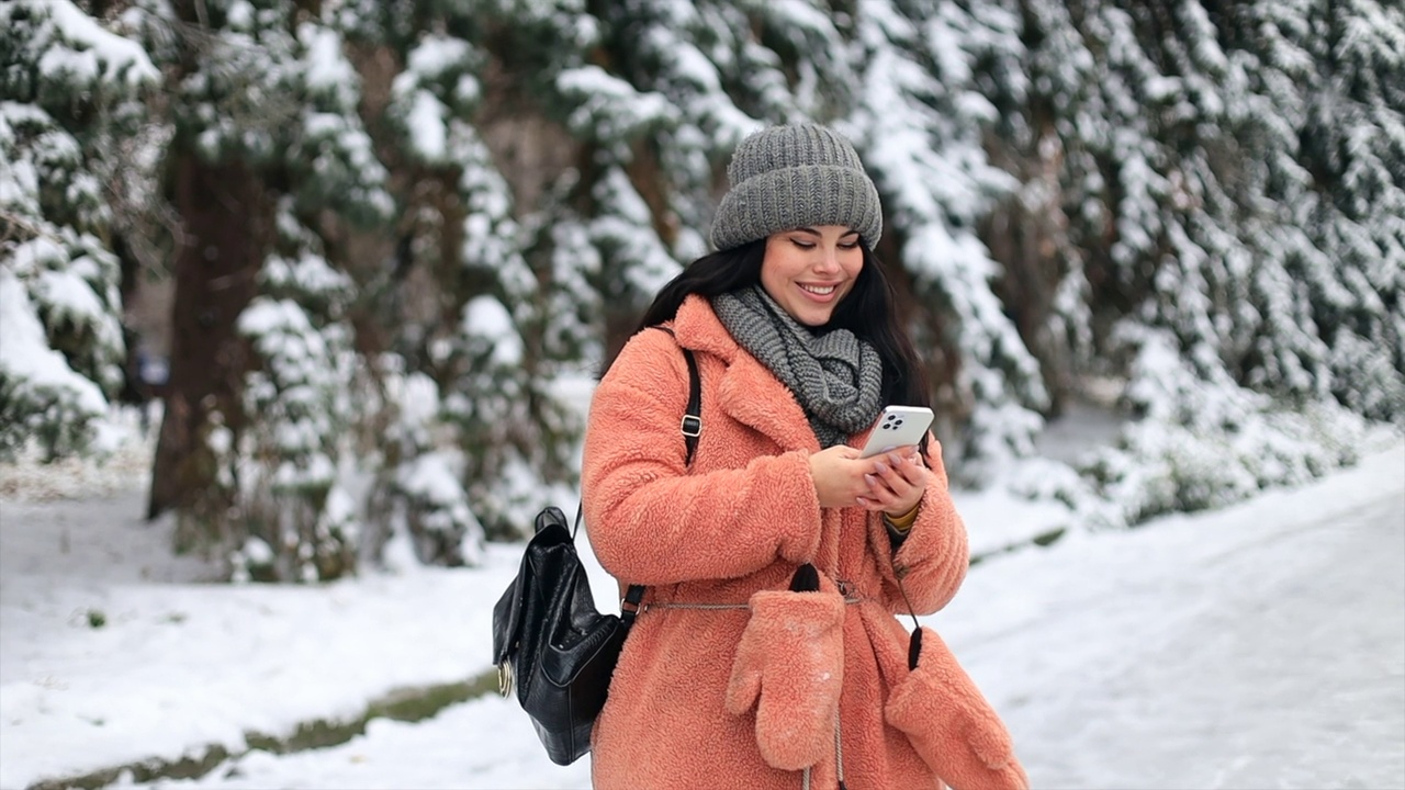 美丽快乐的学生女孩站在冬天的雪地公园在寒冷的天气滚动短信聊天用手机和微笑。视频下载