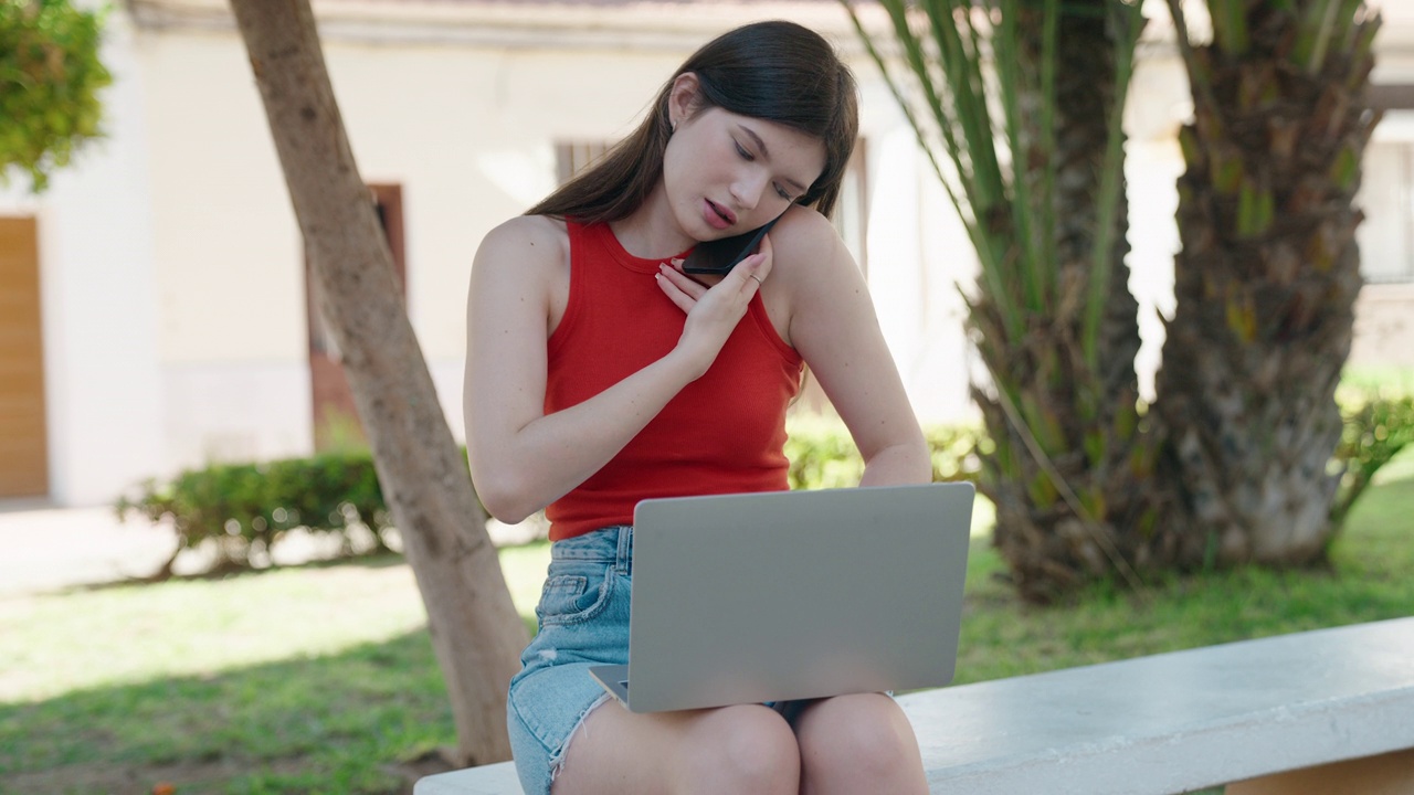 年轻的白人女子坐在公园的长椅上用笔记本电脑用智能手机聊天视频素材