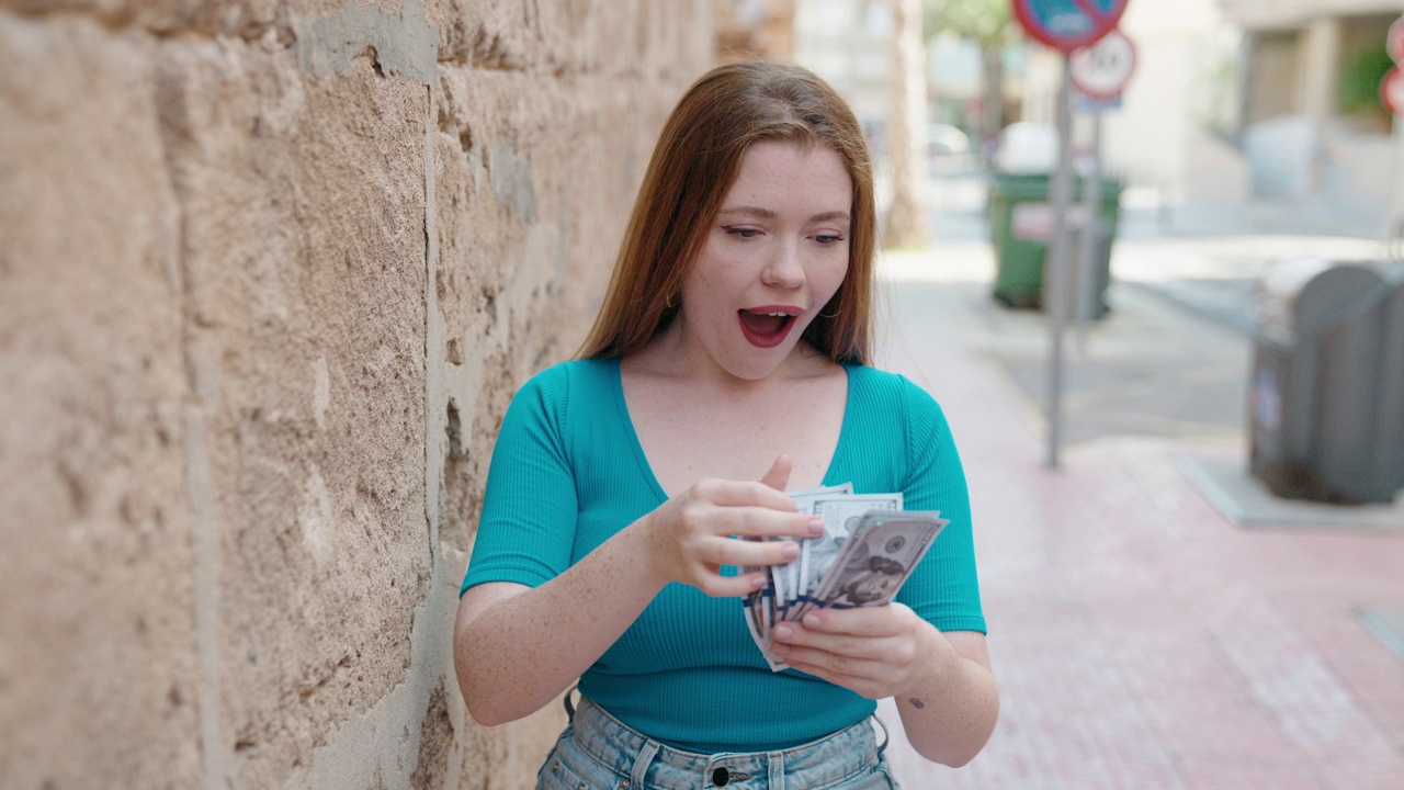 年轻的红头发女人微笑自信地在街上数美元视频素材