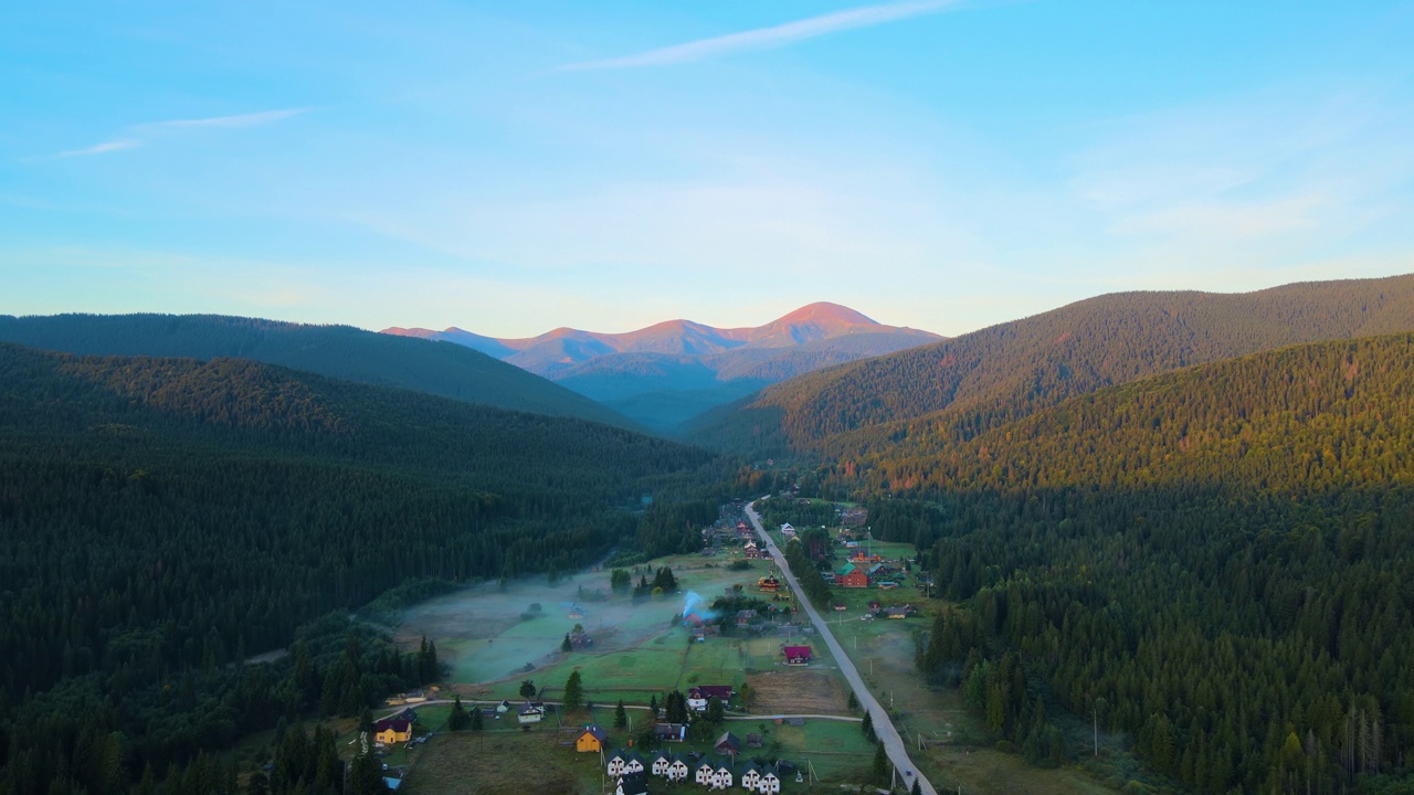 傍晚雾蒙蒙的鸟瞰图，在明亮的夕阳下，在高高的山峰之间的小山村的房子和深色的松林。黄昏时分野山林地的迷人景色视频素材