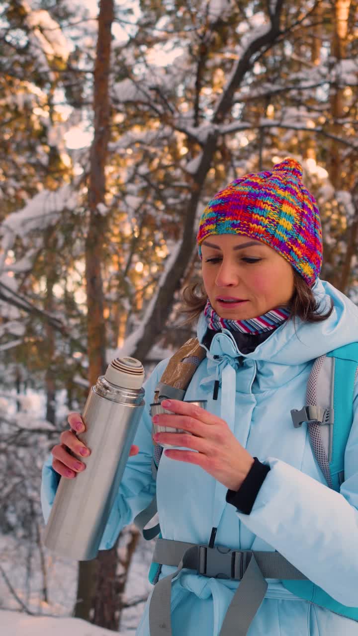 垂直视频显示，一名女子在冬天的雪林里徒步旅行时用保温杯喝着热茶。视频素材