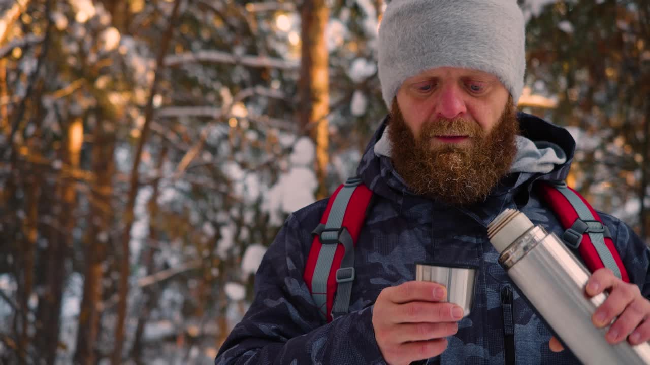 一名中年男子在冬季白雪皑皑的森林里徒步旅行时喝茶的视频。视频素材