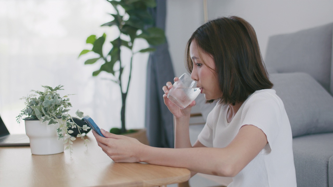 年轻漂亮的亚洲女子坐在家里的客厅里用智能手机发短信，用饮水杯喝水。视频素材