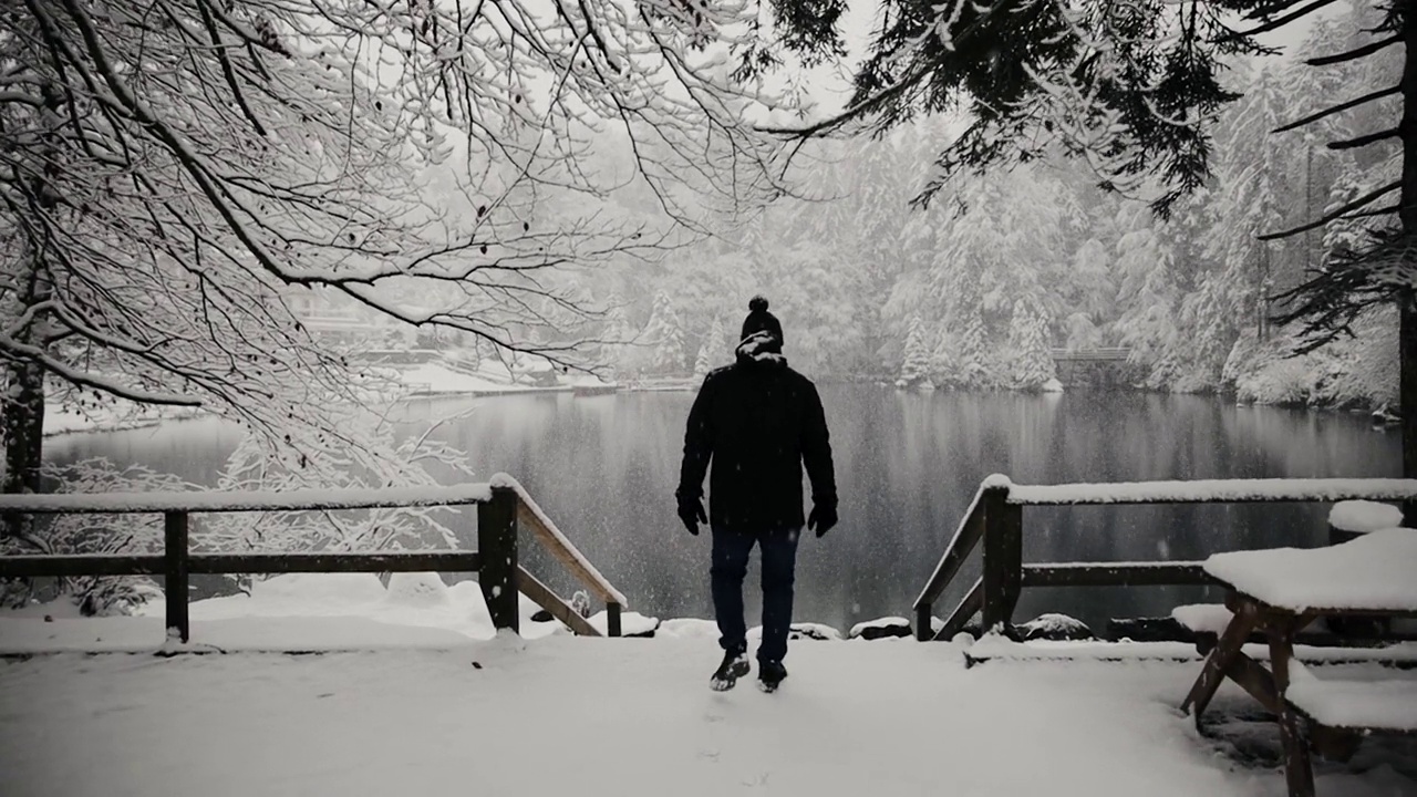 史诗般的镜头，一个孤独的男人走向部分冻结的瑞士山湖。在冬天下雪的时候。男游客穿着保暖的衣服。替身拍摄视频素材