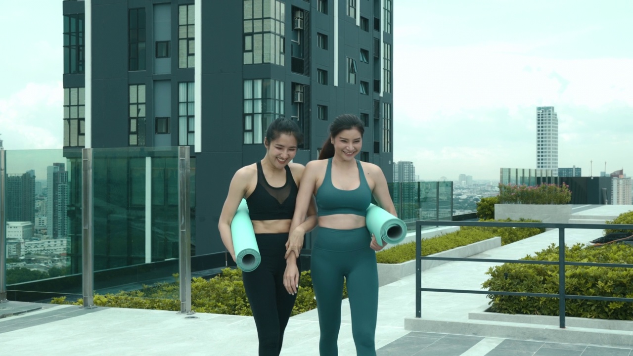 身穿运动服的两名亚洲年轻女性正拿着瑜伽垫，准备在楼顶进行瑜伽练习。视频素材