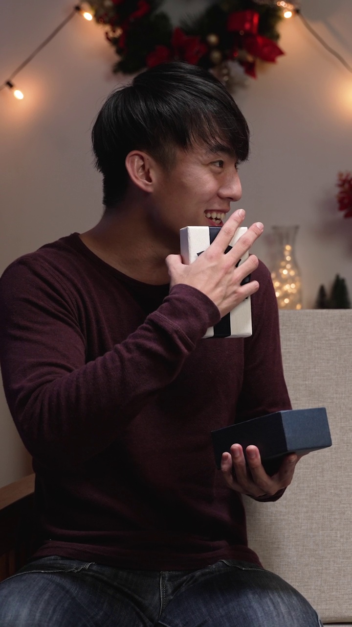 竖屏:亚洲男子惊讶于女友的礼物。一名女子坐在沙发上给震惊的男友送礼物。视频素材