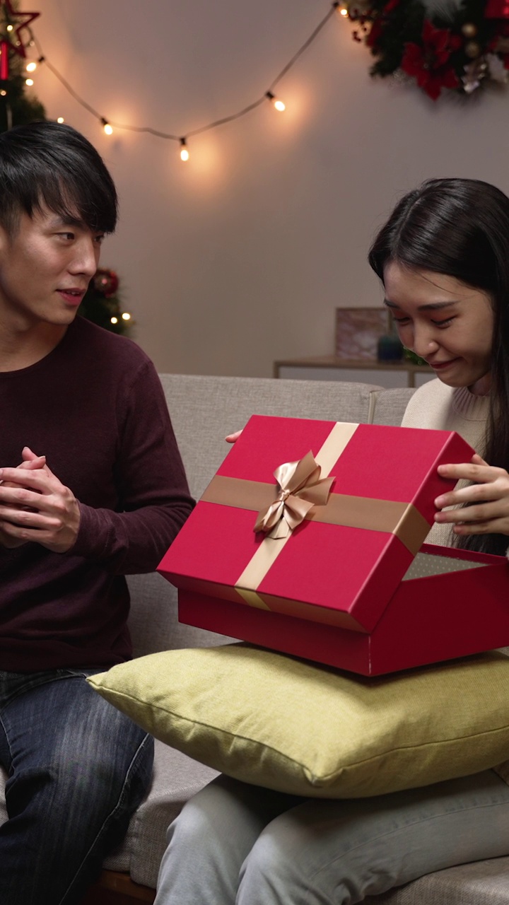 竖屏:平安夜，一对夫妇在家里庆祝。漂亮的女孩满意地收到她男朋友的礼盒坐在家里的沙发上。男人在圣诞节送礼物给妻子。视频素材