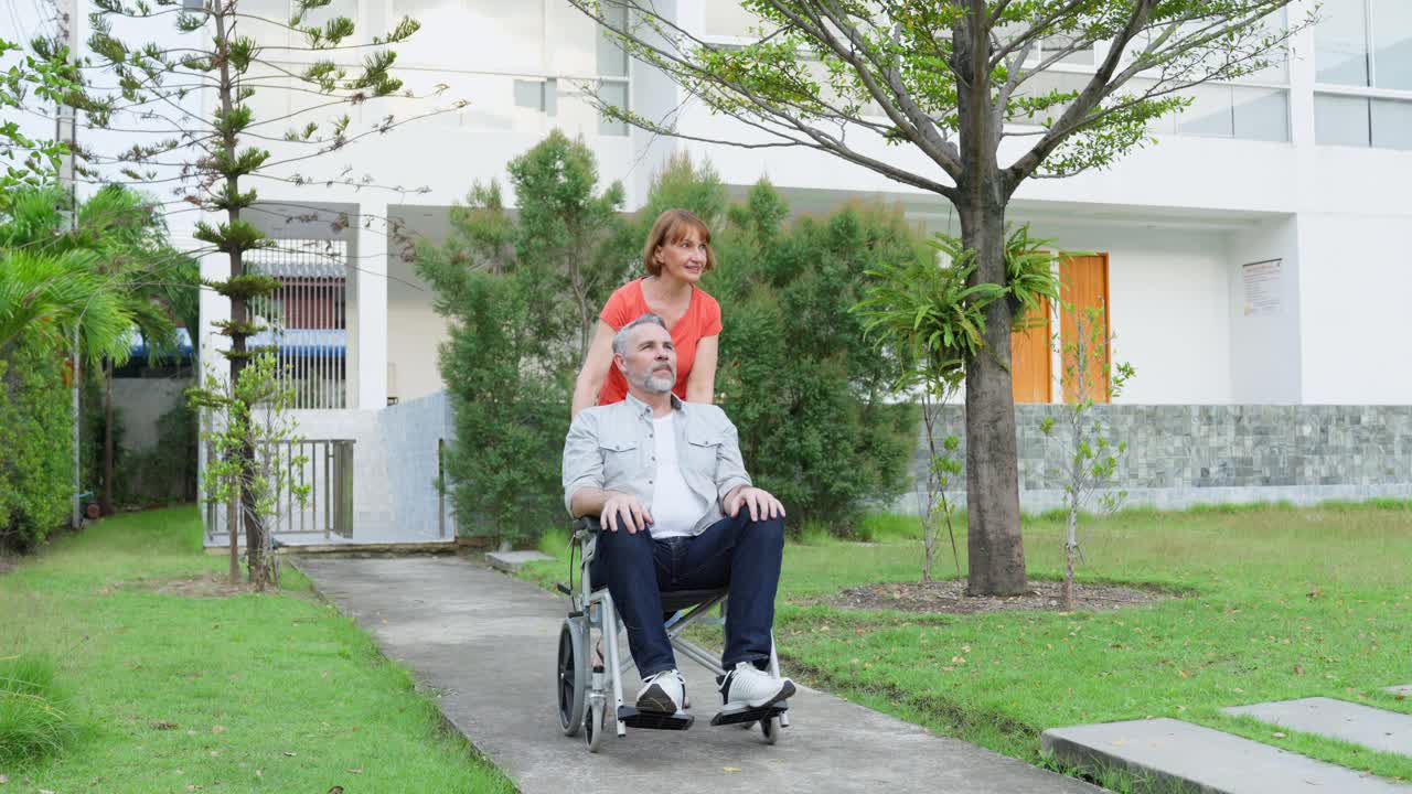 白人老年妇女在室外花园用轮椅支持丈夫。有魅力的年长成熟的妻子照顾和老人病人散步，一起在公园做物理治疗健康。视频素材