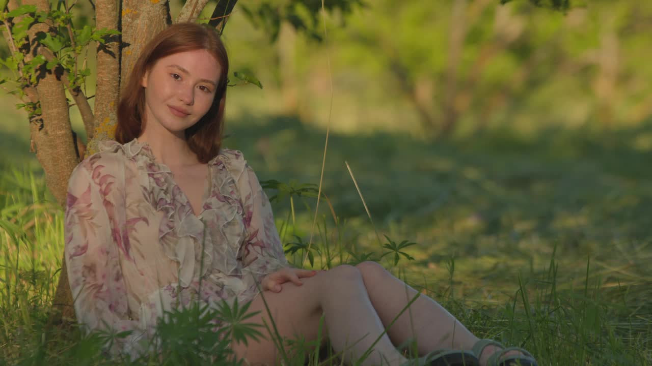 美丽的年轻女孩坐在绿色草地下的树在夏天的国家。自然美景。手持视频视频素材