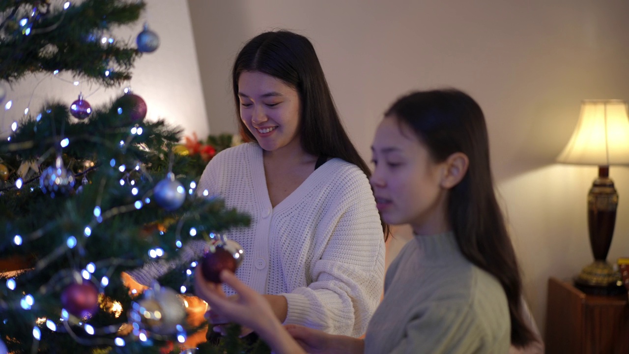 迷人的年轻亚洲女子装饰圣诞树与朋友谈笑。积极美丽的女士们在客厅里享受新年前夜的准备工作。视频素材