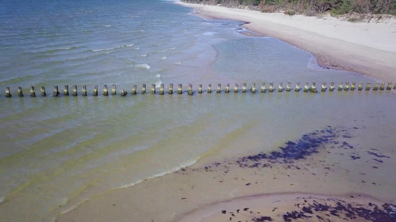 阳光明媚的日子，波罗的海海岸的鸟瞰图，旧的木码头，被海浪破坏的白色沙滩海岸线，海岸侵蚀，气候变化，广角无人机摄影车左移拍摄视频素材