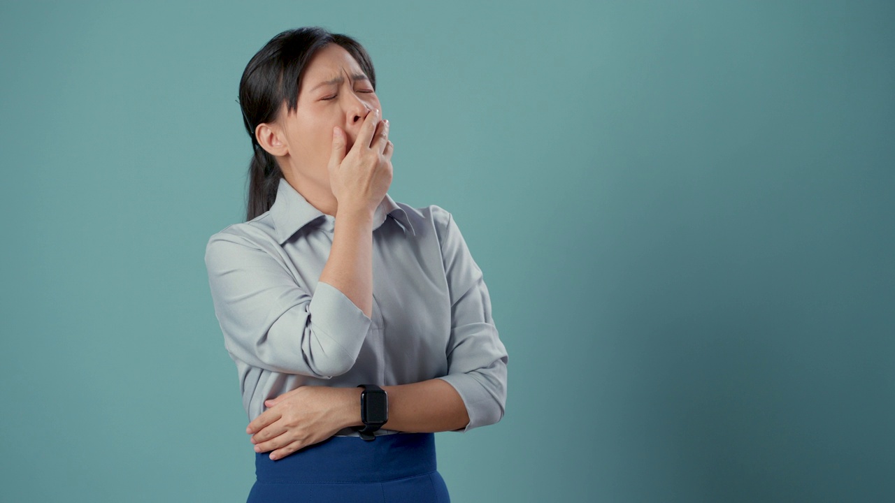 亚洲女性感到疲倦，哈欠连天，孤立地站在蓝色背景下做睡觉的姿势。视频素材