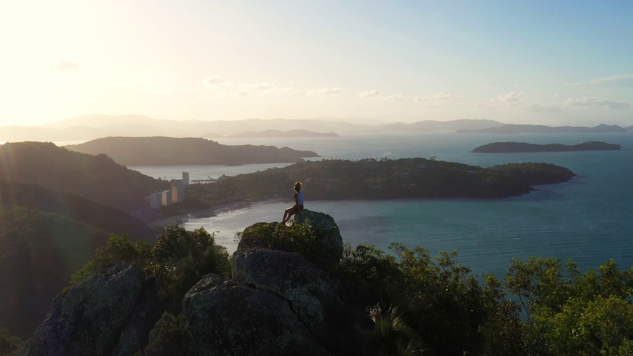 年轻的女性徒步旅行者坐在山顶上，可以看到热带岛屿和蓝色的海洋视频素材