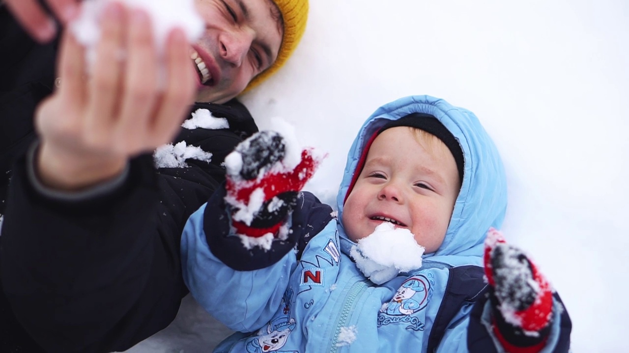 一个冬天的周末，一个快乐的童年。爸爸和男孩躺在雪地里玩雪。冬天里快乐的孩子。雪飘在孩子的脸上。视频下载