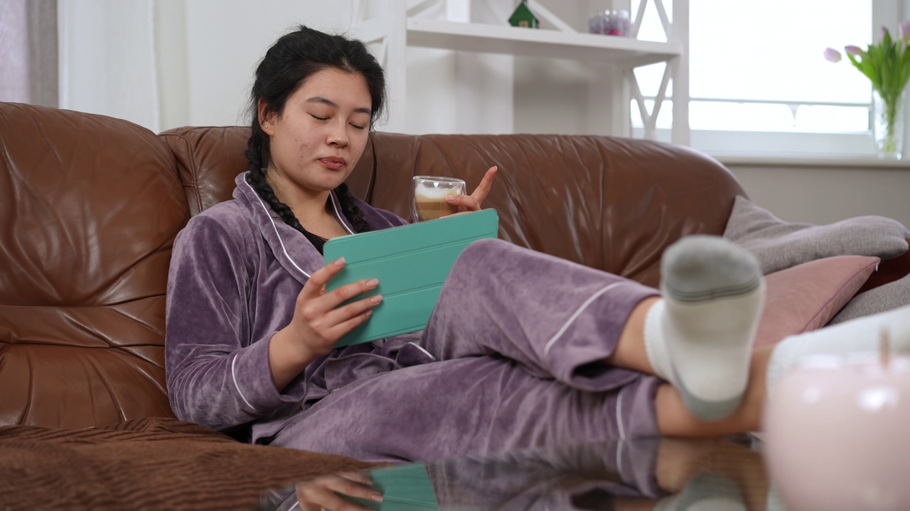 轻松快乐的亚洲年轻女子坐在沙发上看网络系列电影喝咖啡的肖像。无忧无虑的美丽女士在睡衣享受周末休闲沙发与数码平板电脑。视频素材