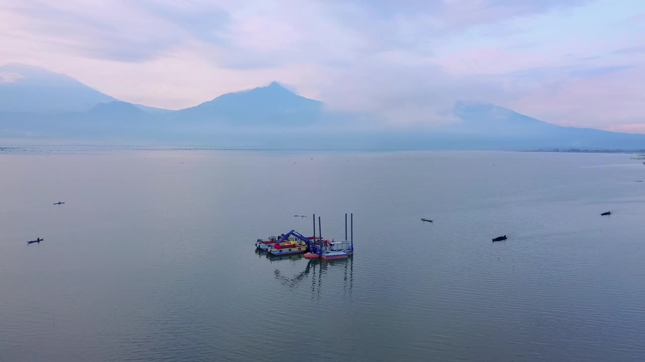 无人机拍摄的挖泥船在湖上清洁和疏浚垃圾-拉瓦湖与山脉的美丽景色为背景，印度尼西亚视频素材