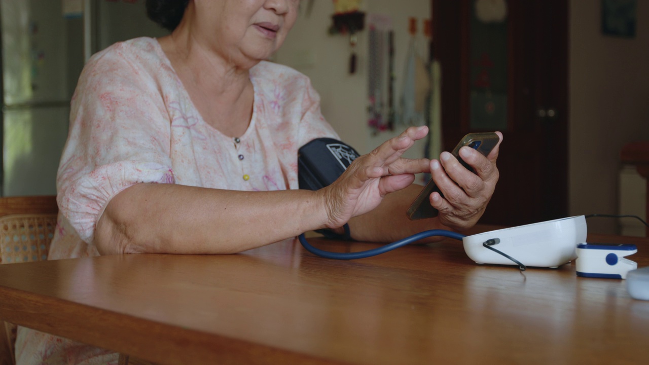 一位年长的亚洲妇女正在家中用数字压力表电眼压计测量血压和心率，并将结果记录在智能手机上。视频下载