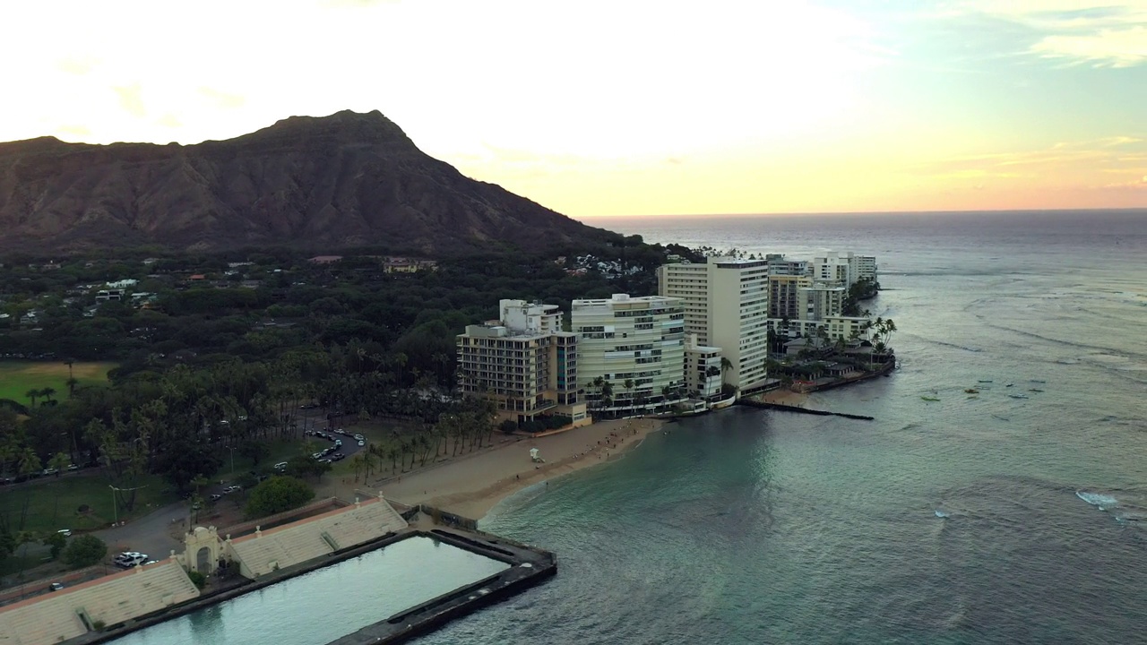 航拍的车辆停在酒店建筑对山，无人机在日落时向后飞越海景-瓦胡岛，夏威夷视频素材