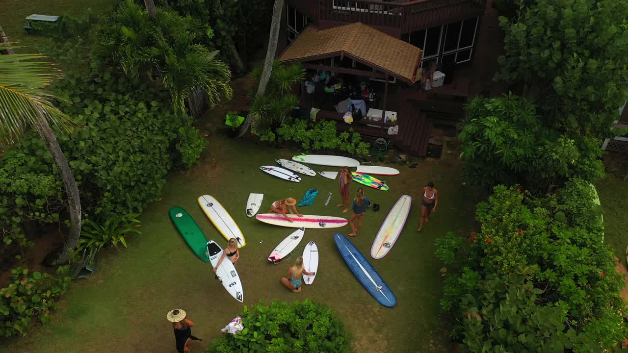 航拍女性朋友在小屋外冲浪板，无人机向后飞过绿色植物——夏威夷瓦胡岛视频素材