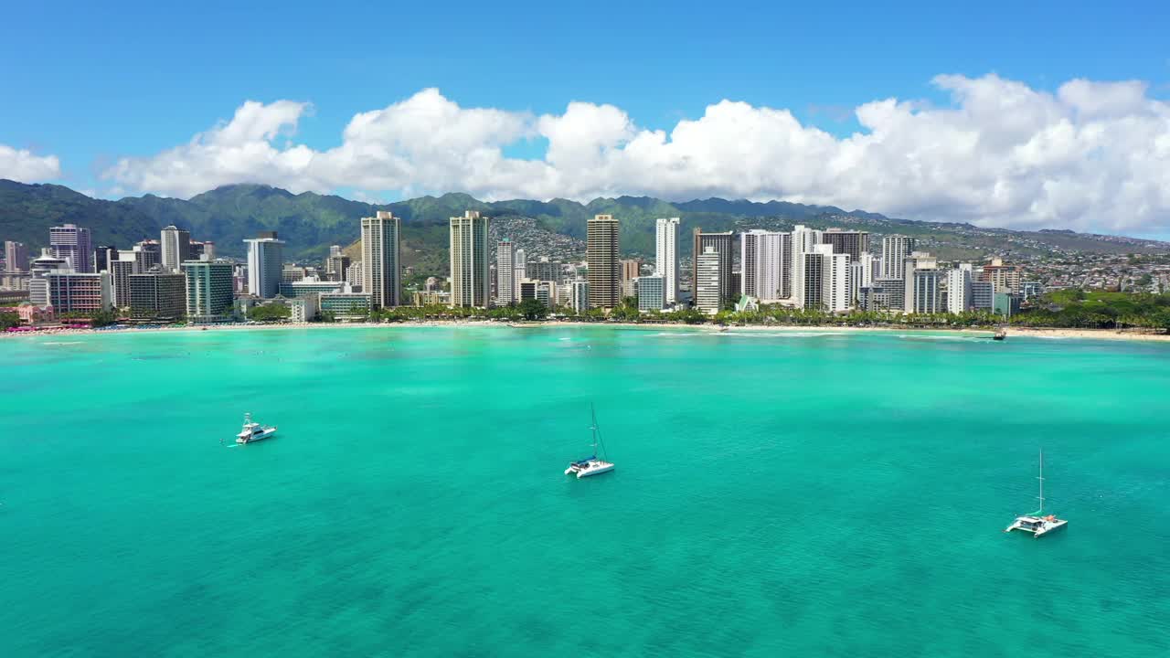 空中向下拍摄的白色帆船漂浮在蓝绿色的大海上，现代城市对多云的天空-瓦胡岛，夏威夷视频素材